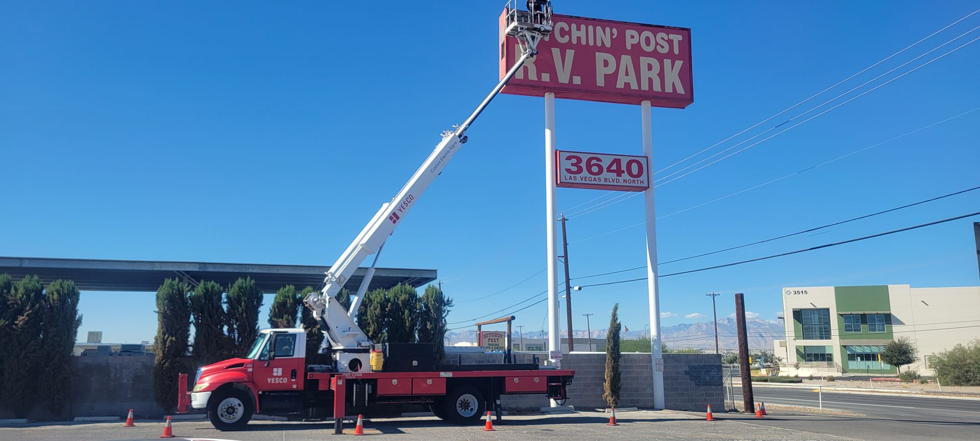 A truck with a lift repairs the Watchin' Post R.V. Park sign, against a blue sky.