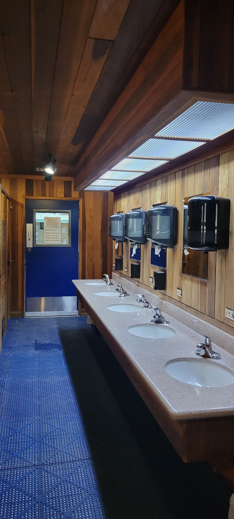 Bathroom with wooden paneling, a row of sinks, blue tile floor, and a blue door.