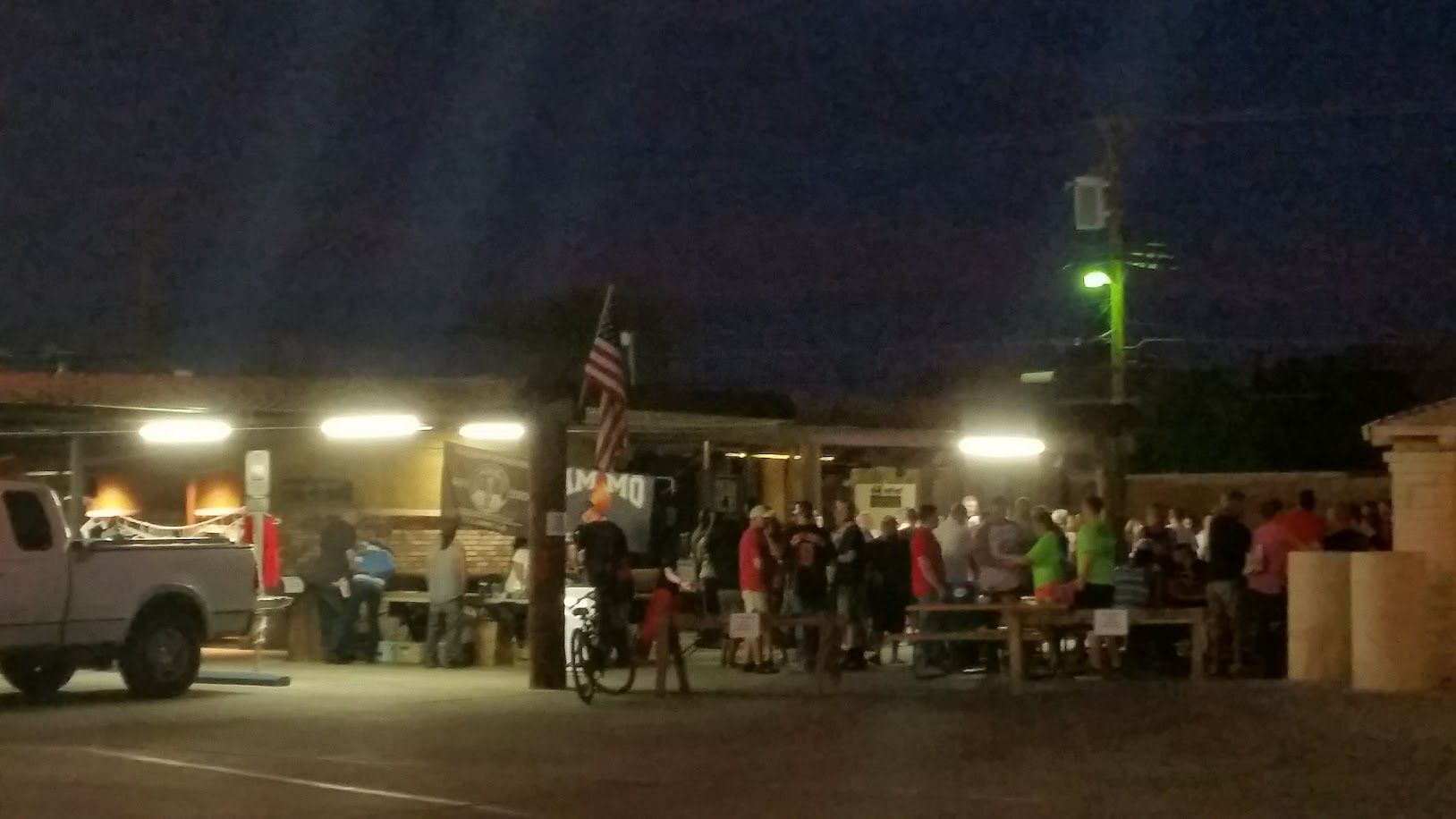 Nighttime gathering outside a building with people, tables, and a truck. American flag visible.