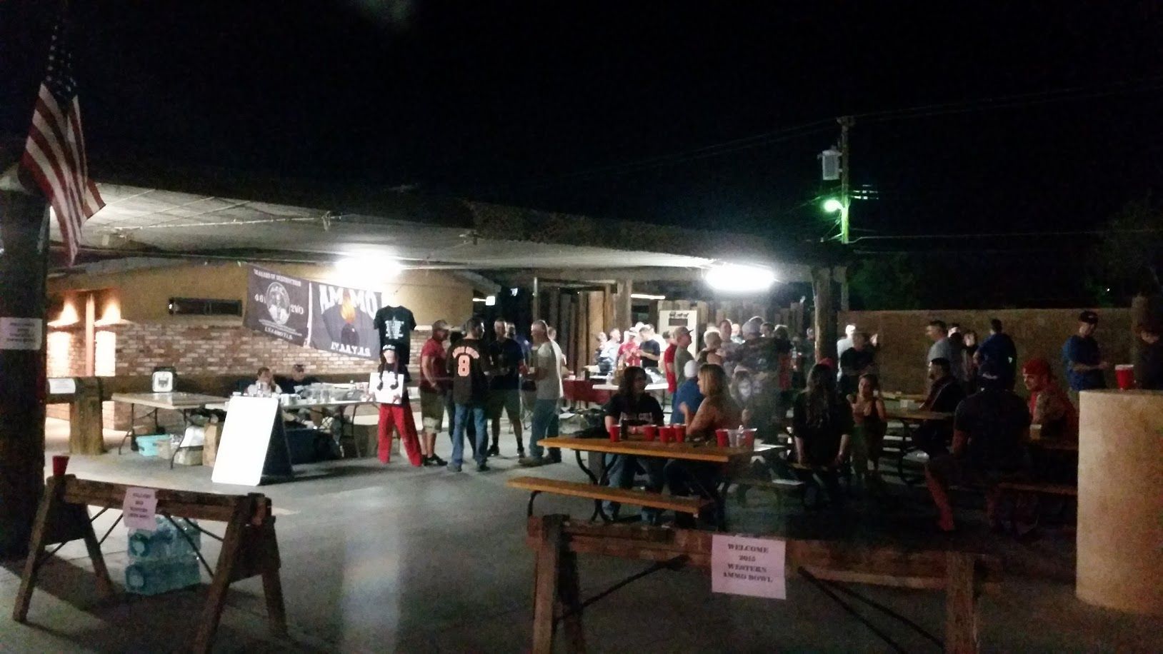 Night scene: outdoor gathering under a covered area; people at tables, American flag visible, lit by overhead lights.
