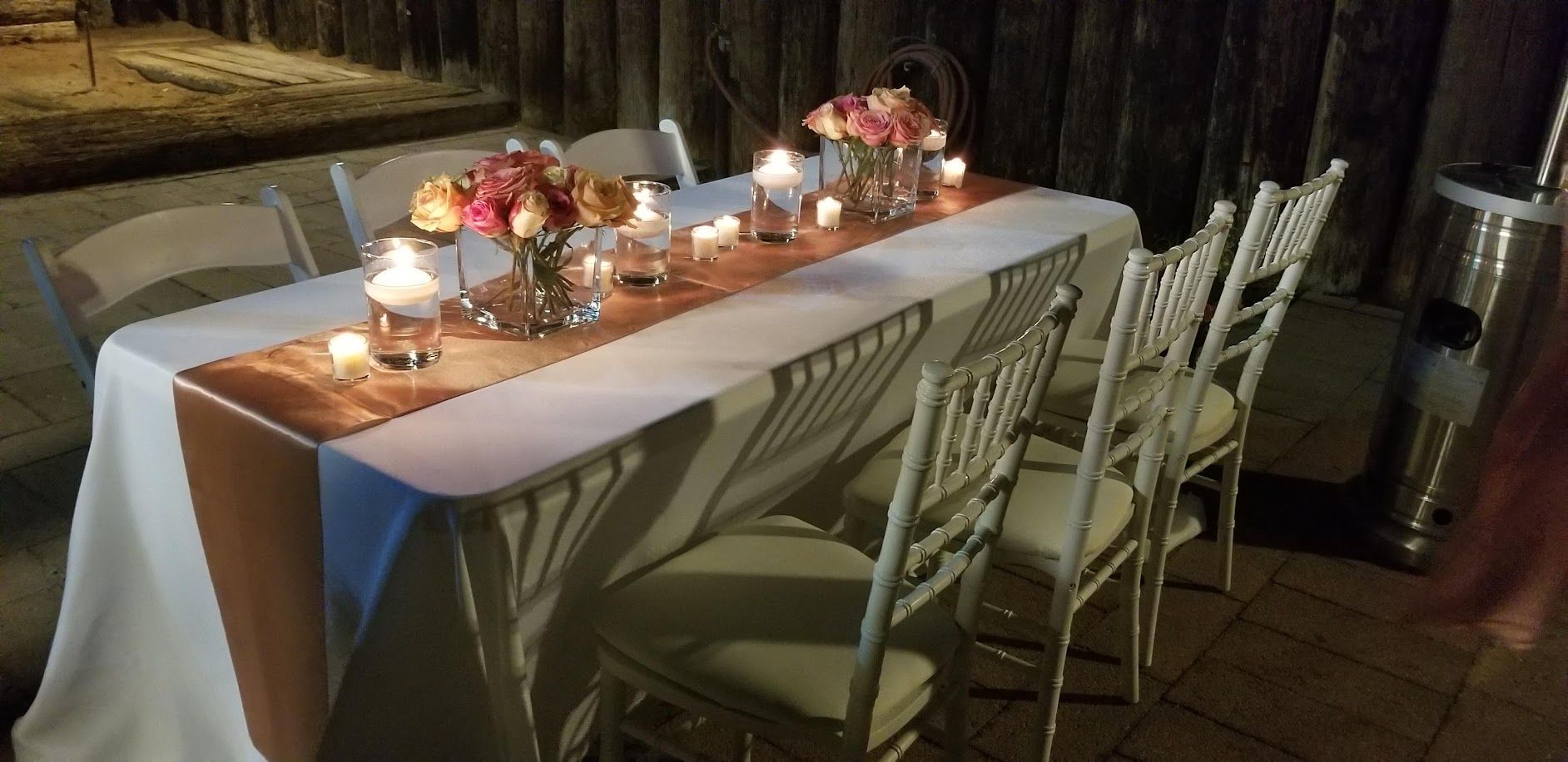 Table set for a party with flowers, candles, and chairs, likely outdoors.