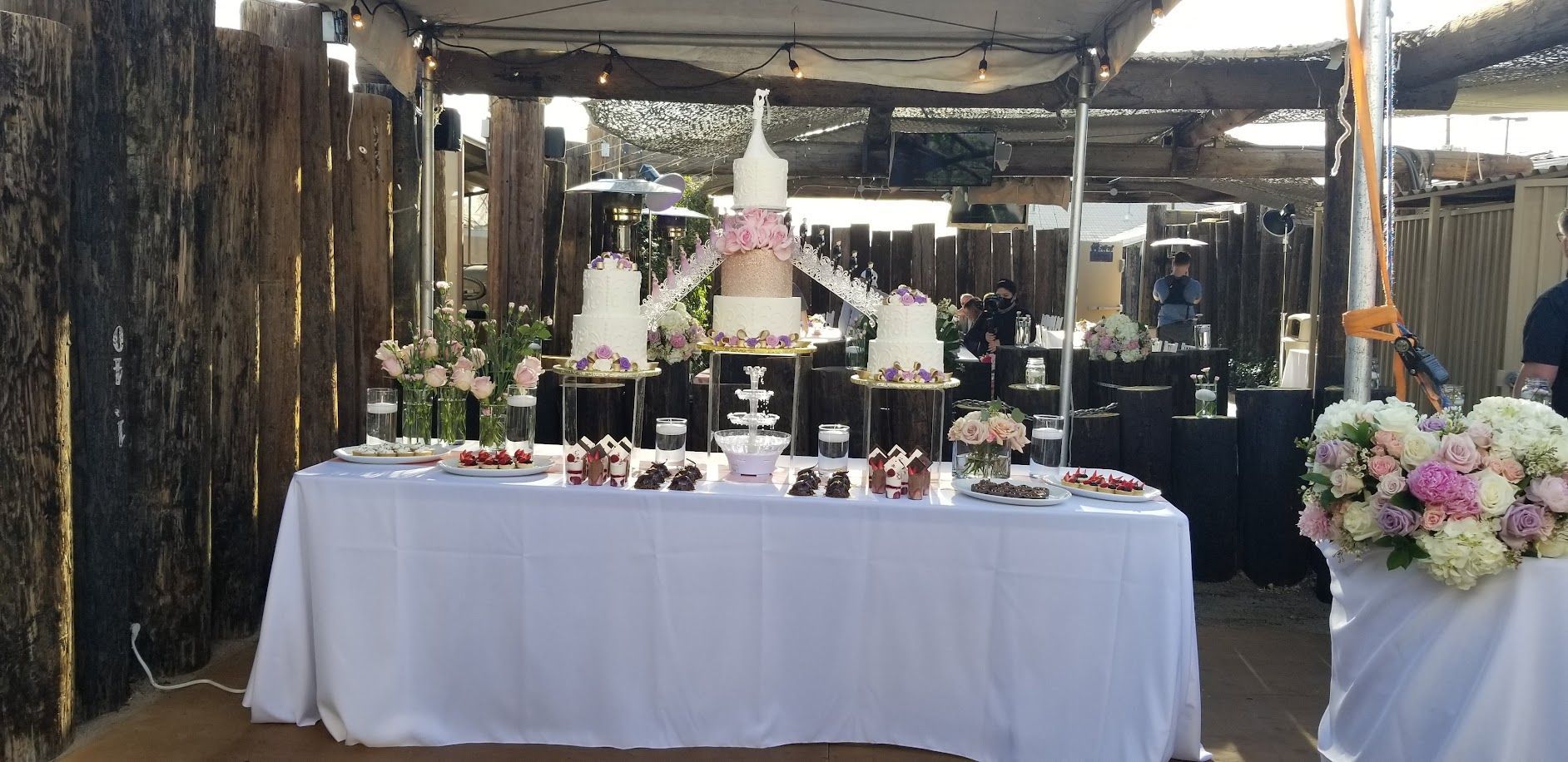 Wedding dessert table with cakes, flowers, and treats. Outdoor setting with rustic wood wall.