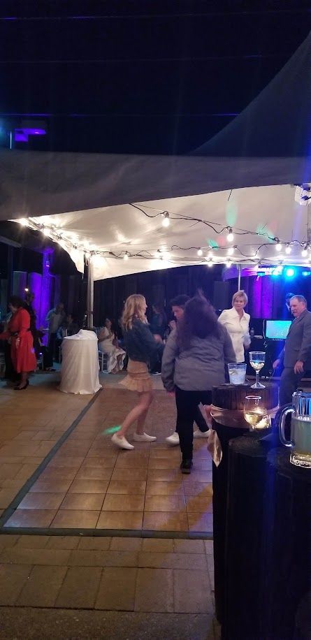 People dancing on a wooden dance floor, under a canopy with string lights. A bar is in the foreground.