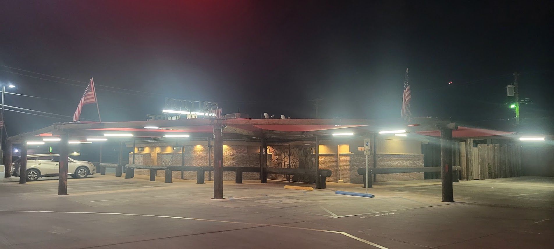 A nighttime shot of a restaurant with a covered entrance, a parked car, and flags.