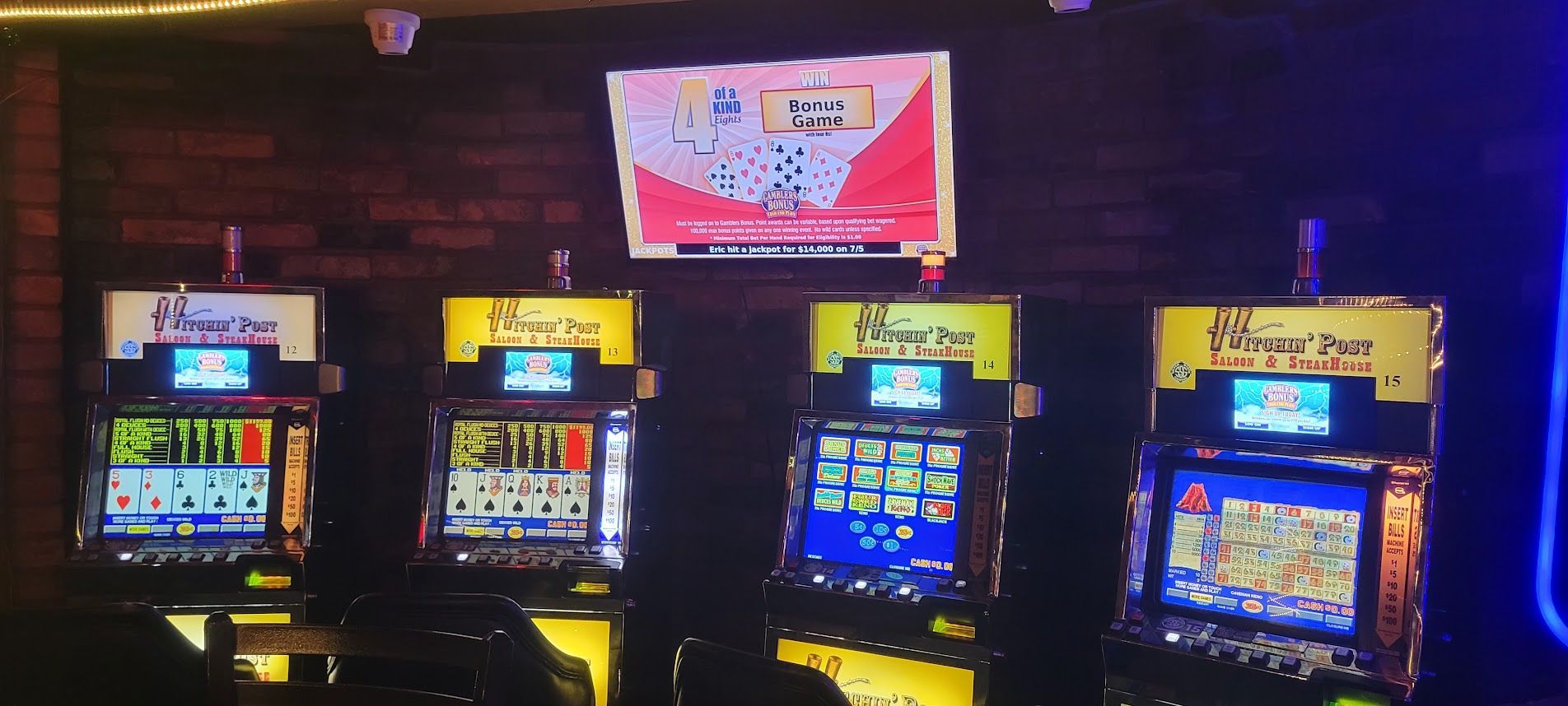 Four slot machines in a casino setting, with a screen displaying information above them.
