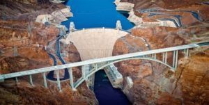 Aerial view of Hoover Dam and Mike O'Callaghan–Pat Tillman Memorial Bridge. The dam is tan, and the bridge is white, both amidst rocky terrain.