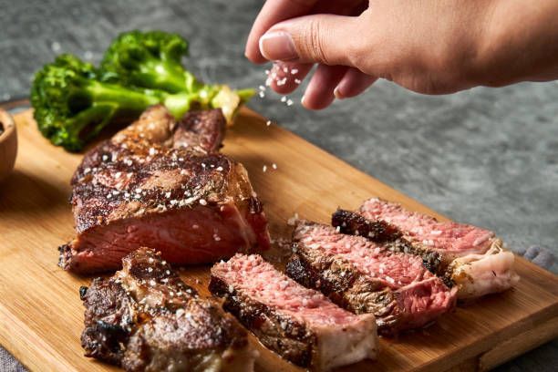 Steak slices on wooden board; hand sprinkling salt. Broccoli sprig in background.