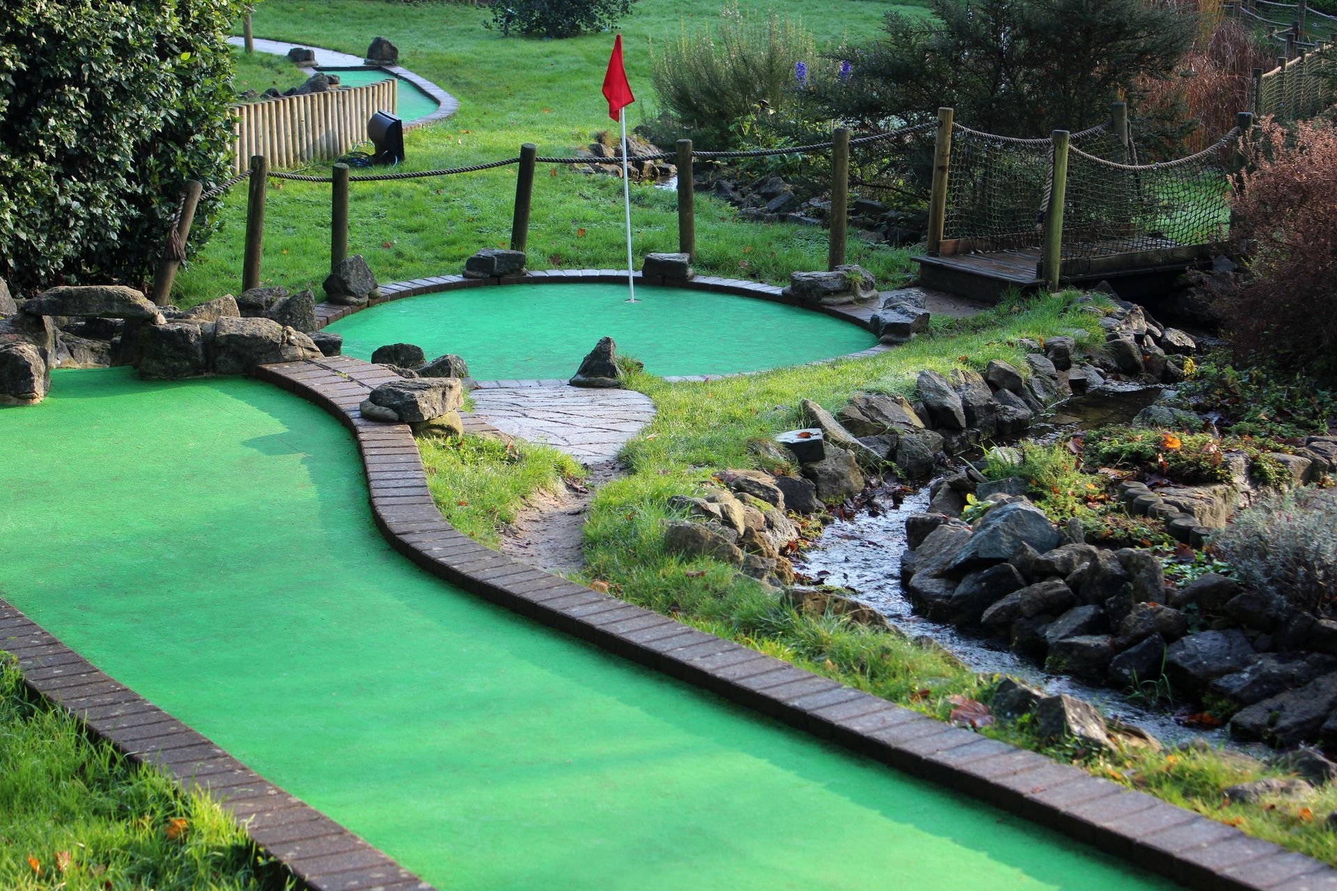 Mini golf course with green turf, bridge, water feature, and red flag.