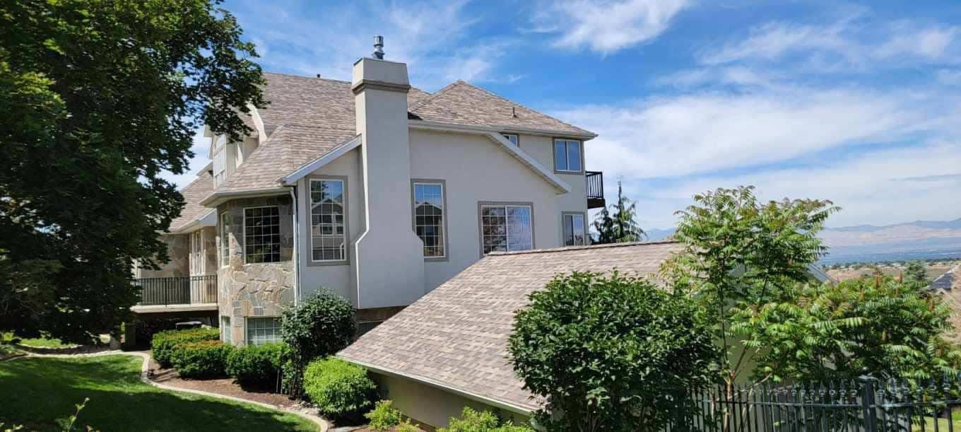 Large house with light colored walls, brown roof, chimney, and surrounding trees. Blue sky.
