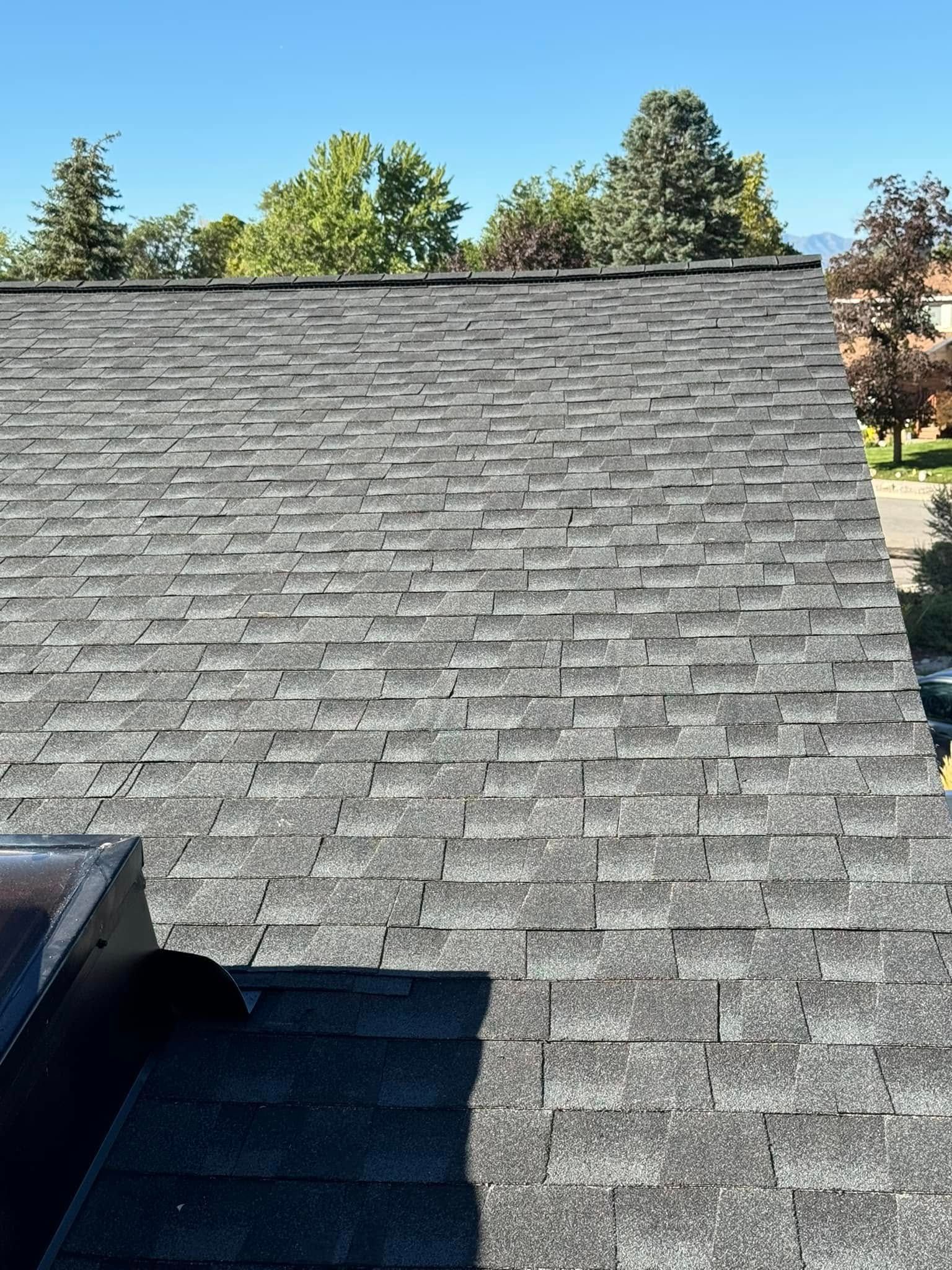 Gray asphalt shingle roof on a sunny day with blue sky and trees in background.