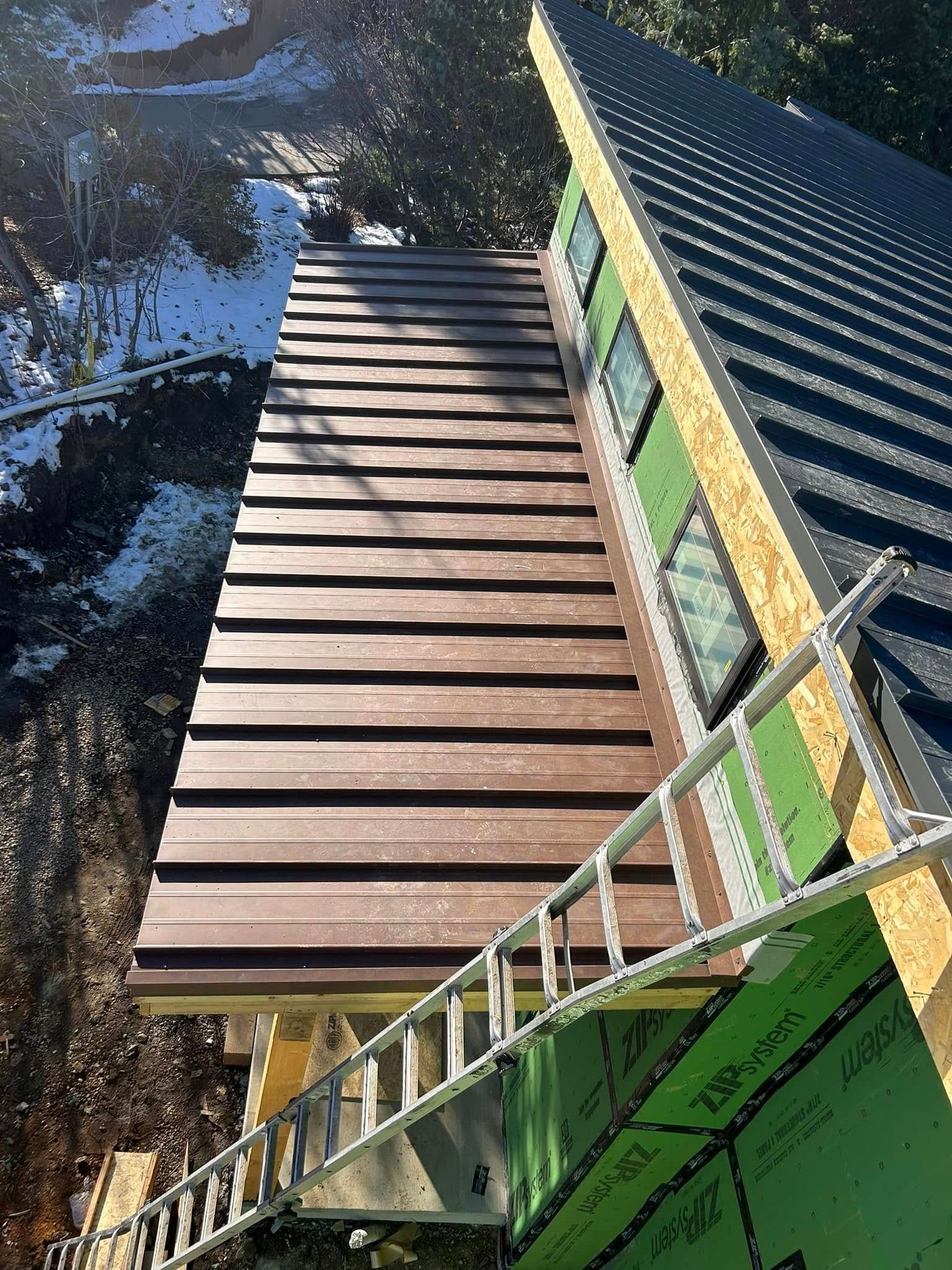 Construction site: brown metal roof, green sheathing, ladder, windows, snowy background.