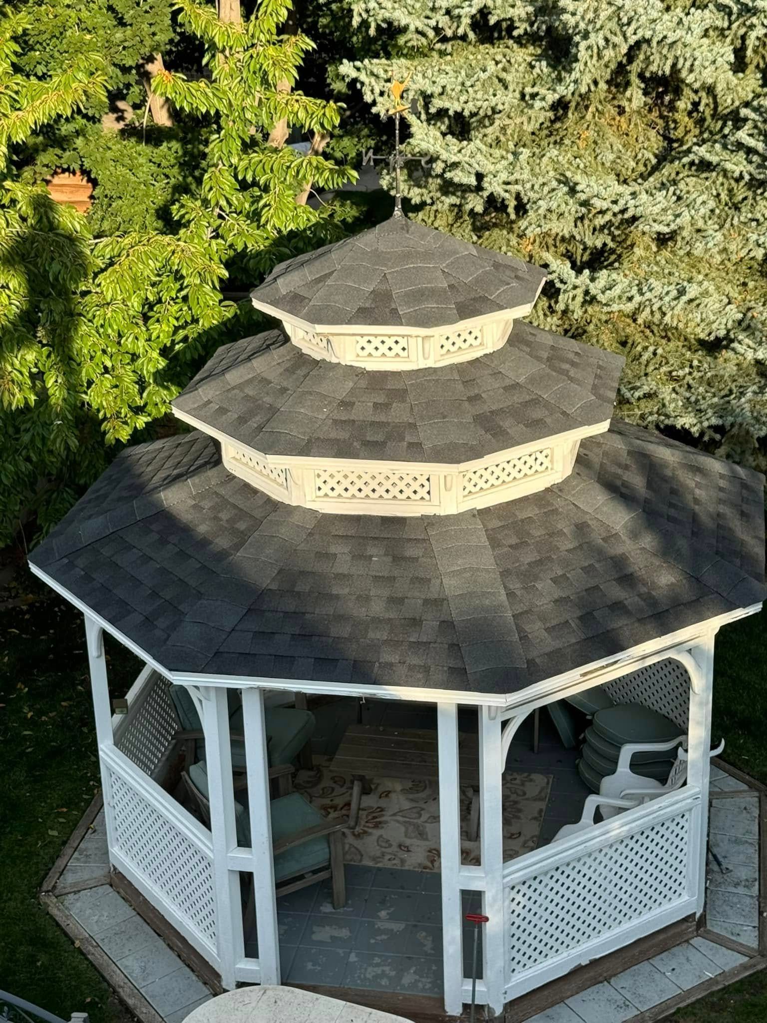 White gazebo with black roof, lattice walls, and seating in a green yard.
