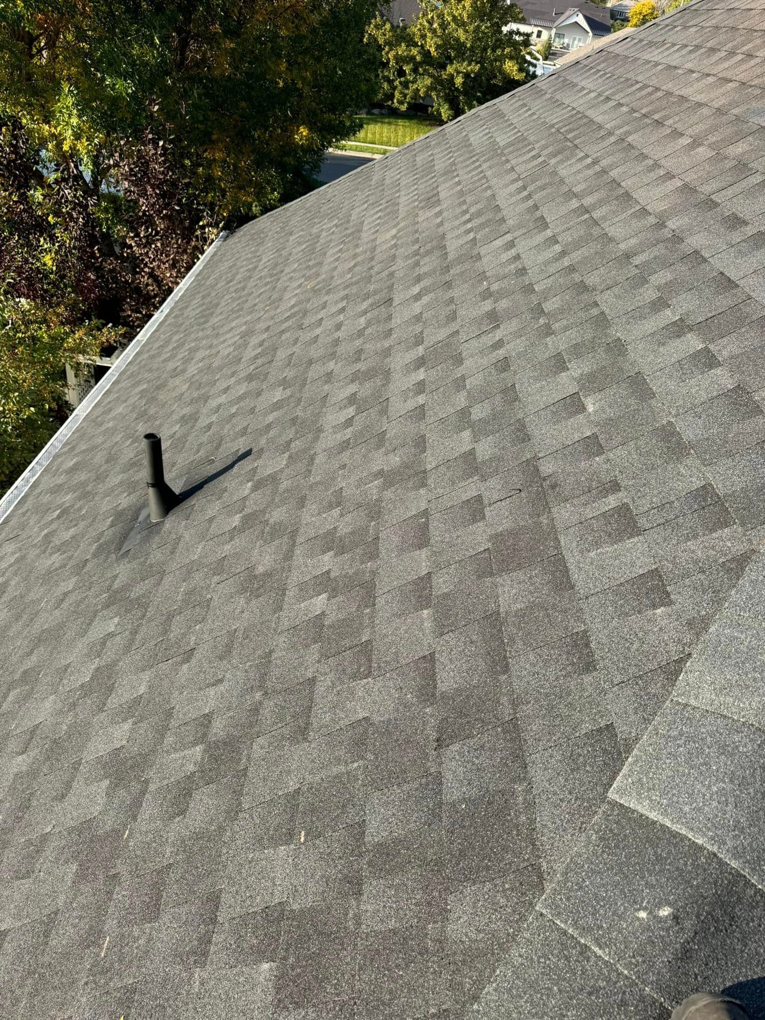 Gray asphalt shingle roof with a vent pipe; sunlight illuminates the shingles and surrounding trees.