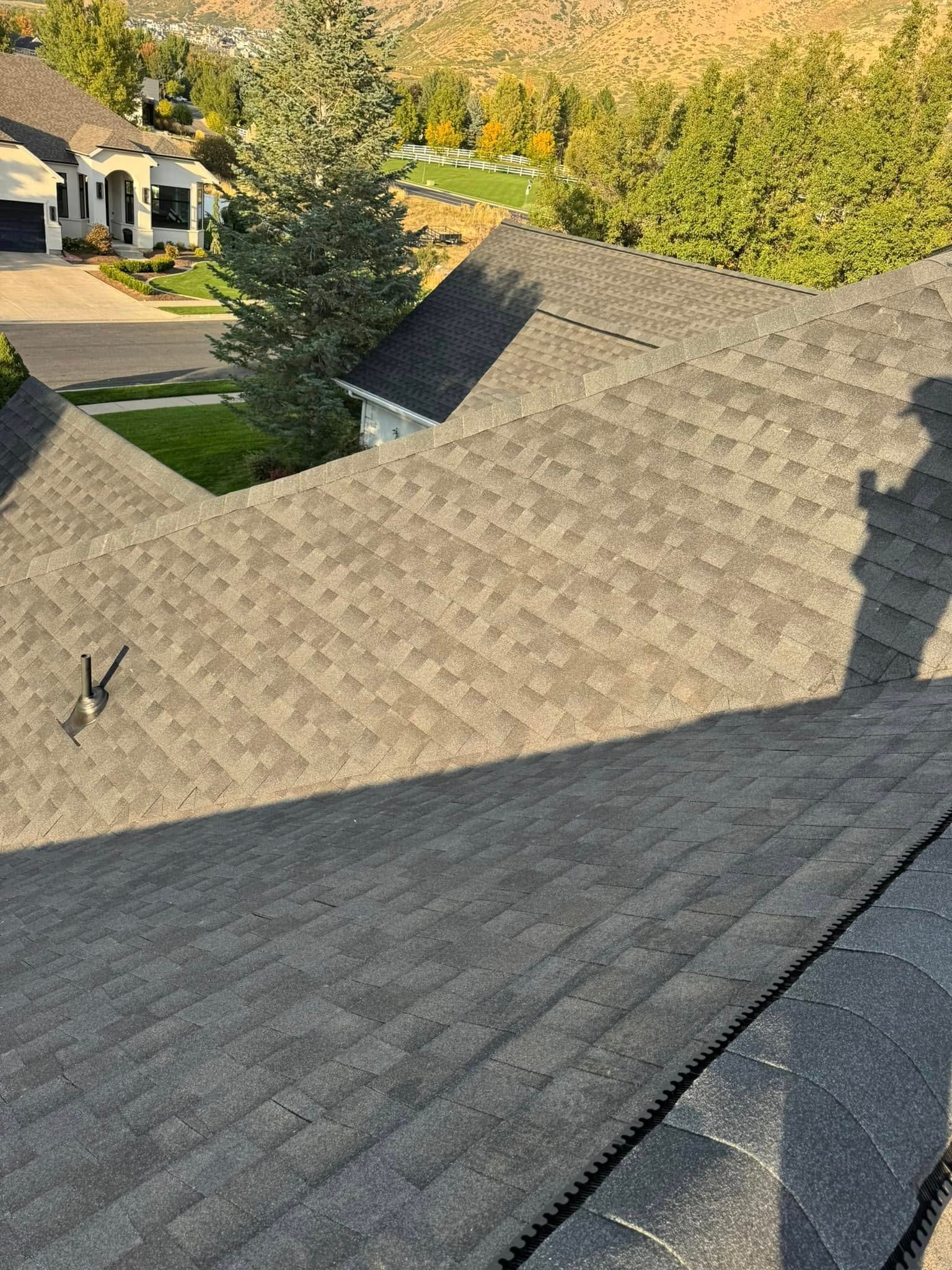 A dark shingle roof with shadows, in a neighborhood with trees and houses.