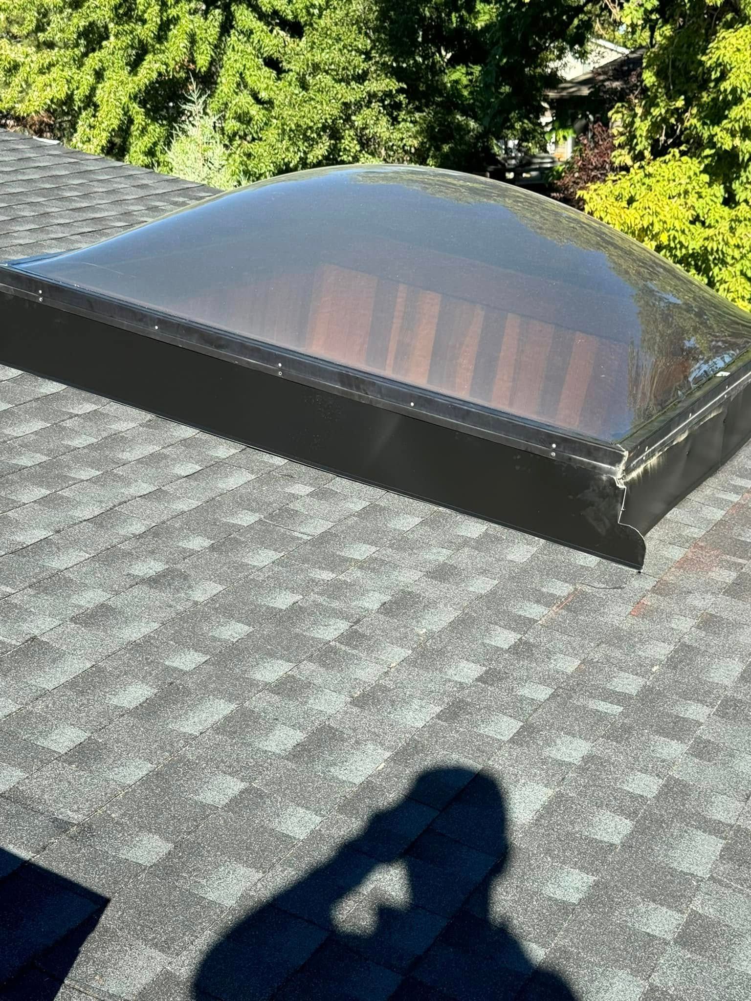 Skylight on a shingled roof. The domed, tinted plastic is in a black frame. Shadow of a person in the foreground.