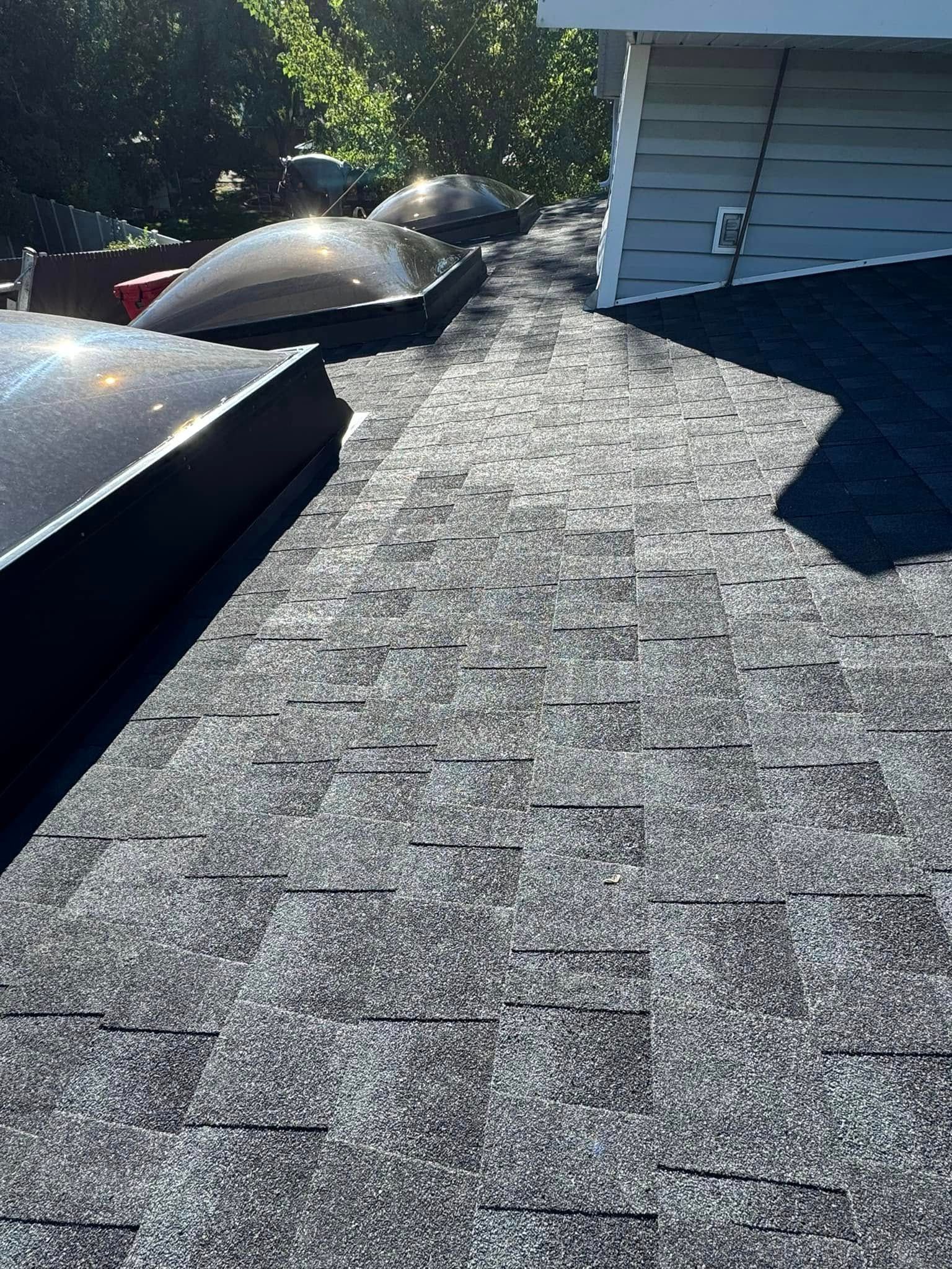 Dark asphalt shingle roof with two skylights, next to a white house wall. Sunny outdoor setting.
