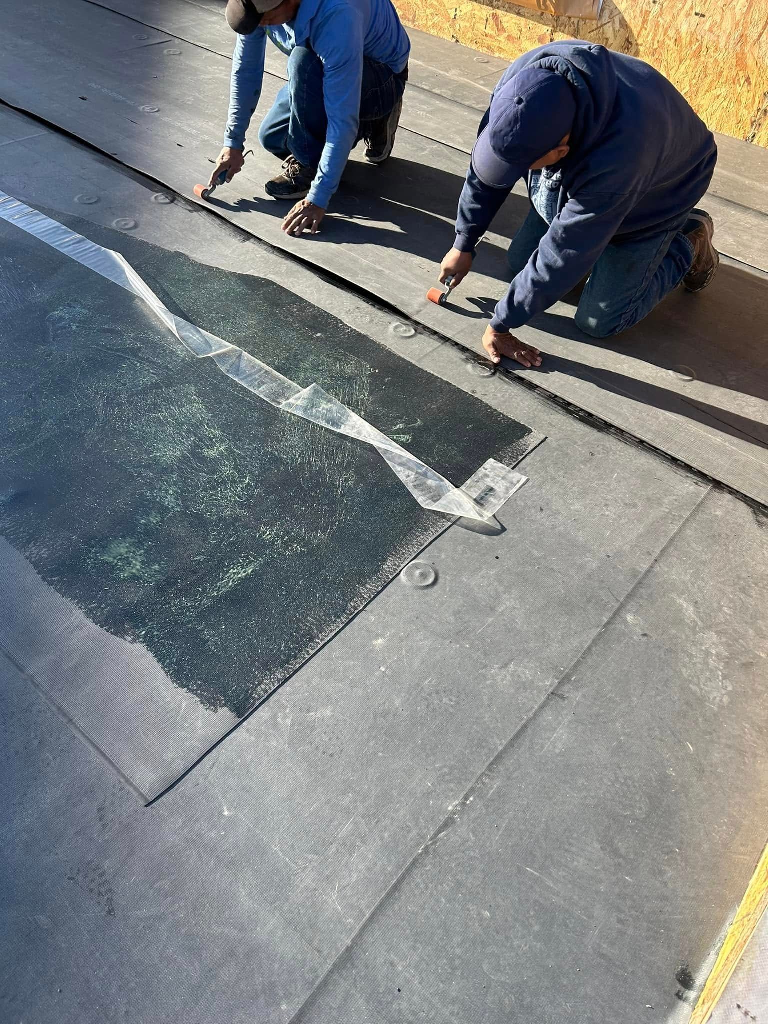 Two men kneeling, installing black roofing material.