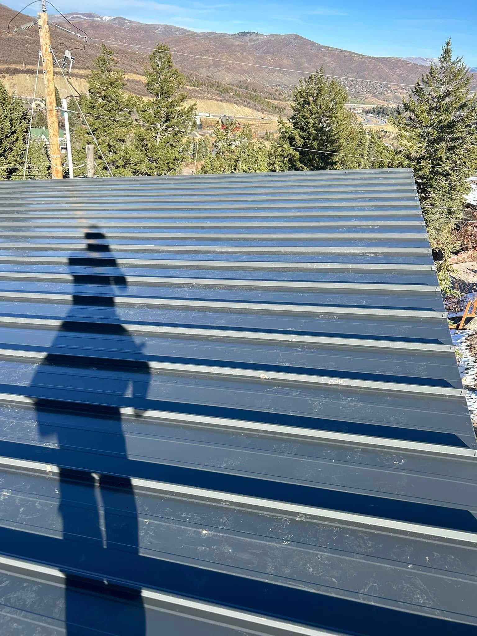 Shadow of a person on a ribbed, metal roof with a mountain backdrop on a sunny day.