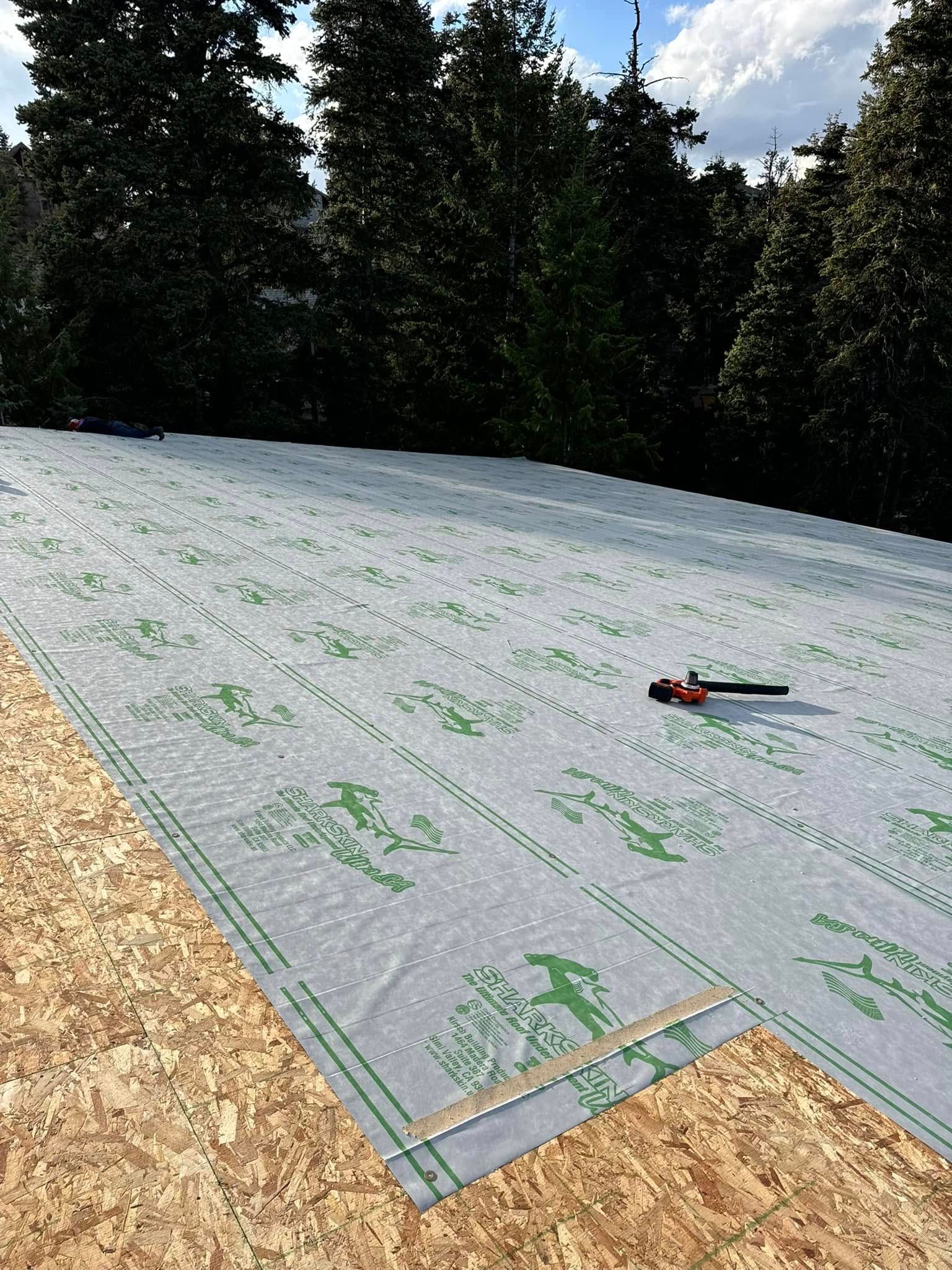 Roofing underlayment being installed on a wooden surface. Green and gray materials against trees and sky.