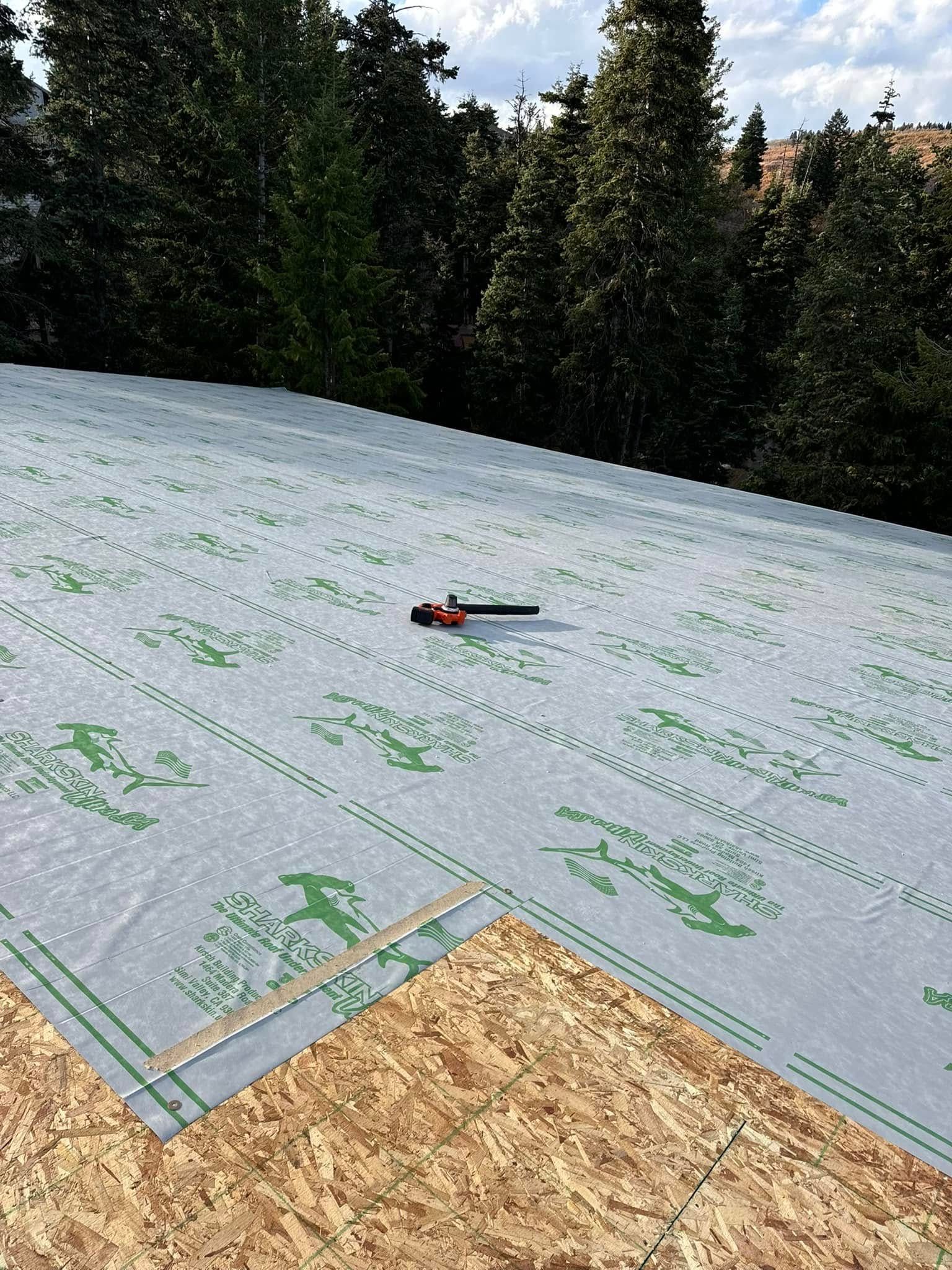Rooftop with sheathing, underlayment, and tools. Green and gray underlayment against a wooded background.