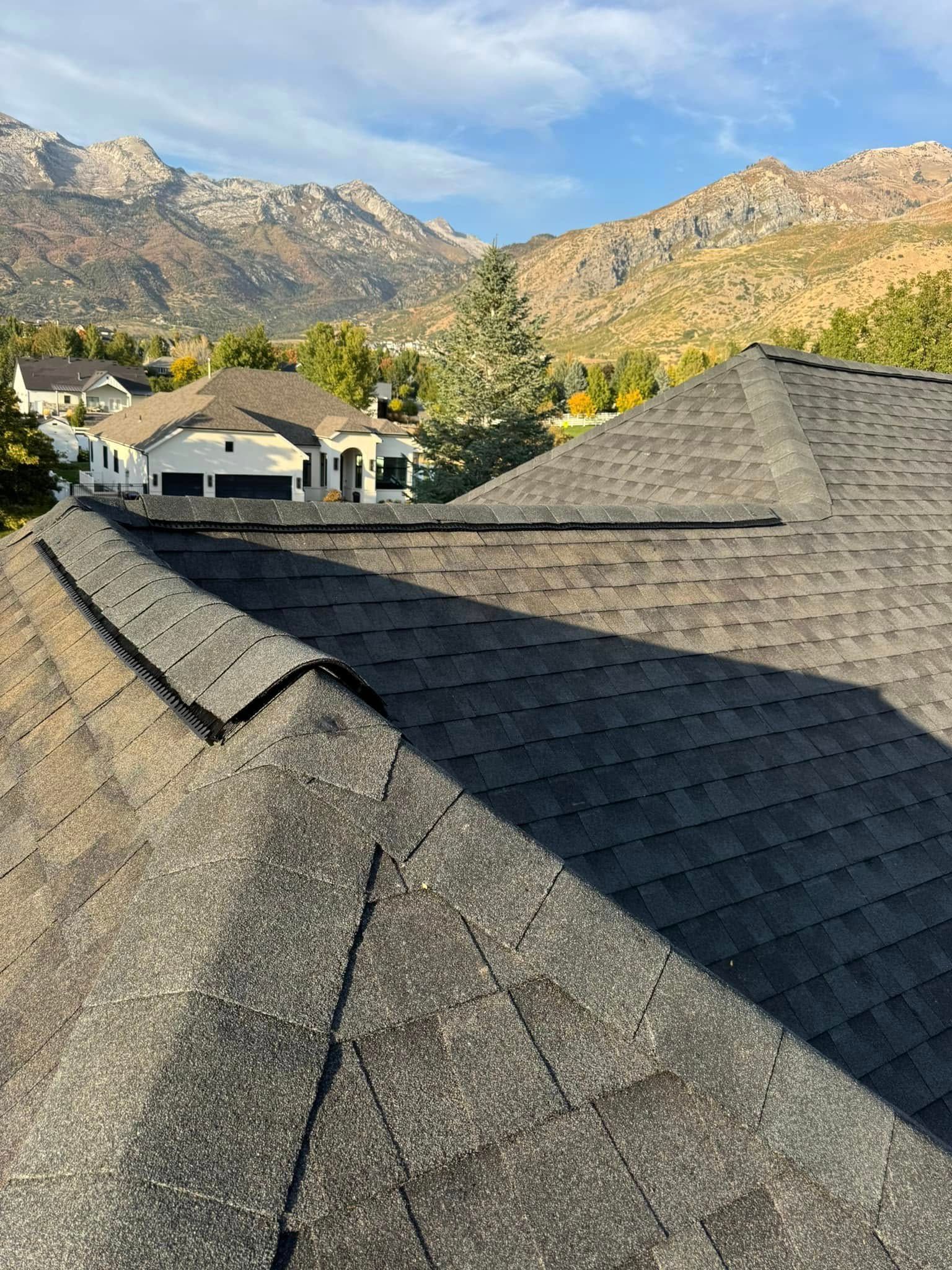 View of a dark shingled roof with mountains and houses in the background under a blue sky.