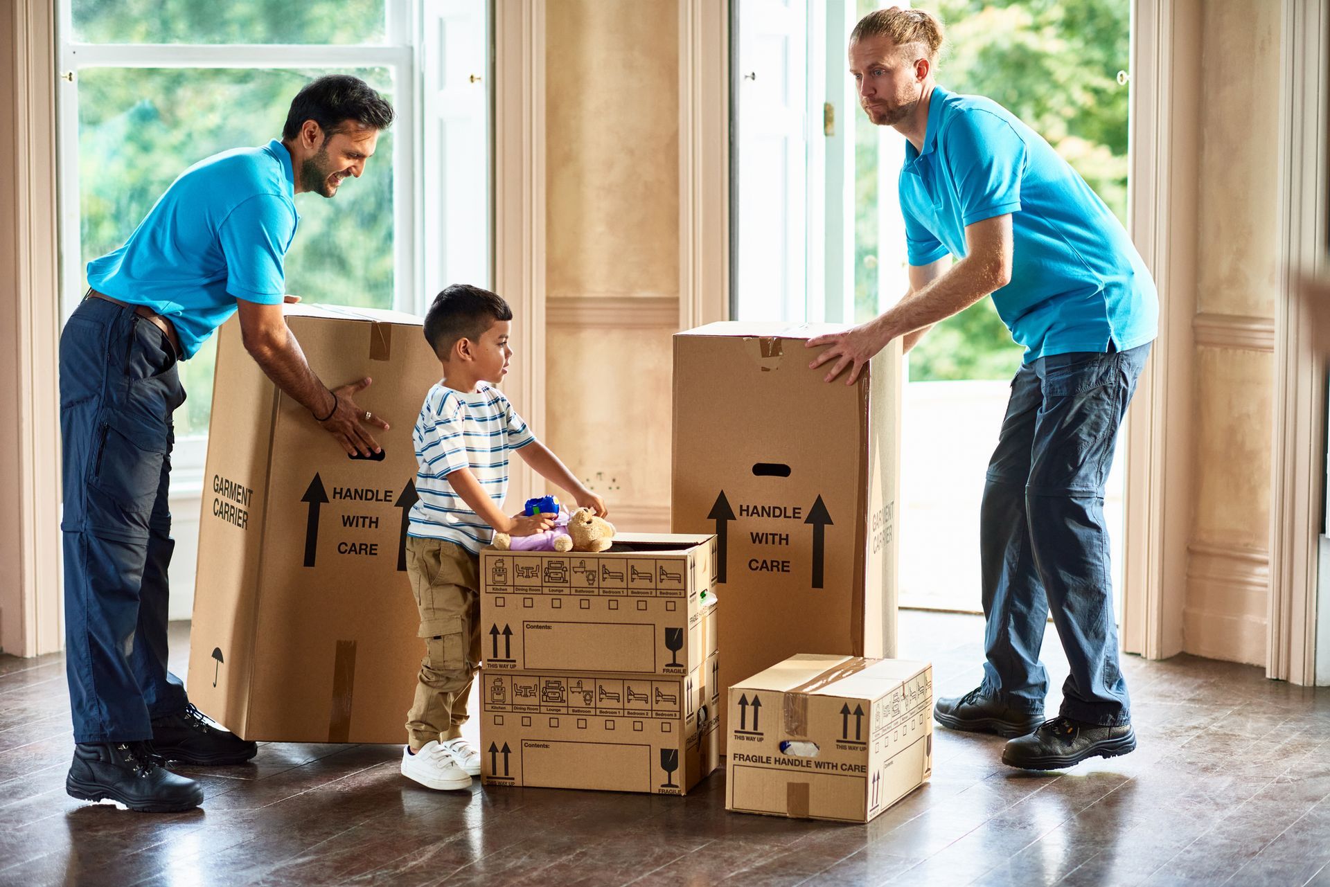 Two movers carry boxes with a child, inside a home.