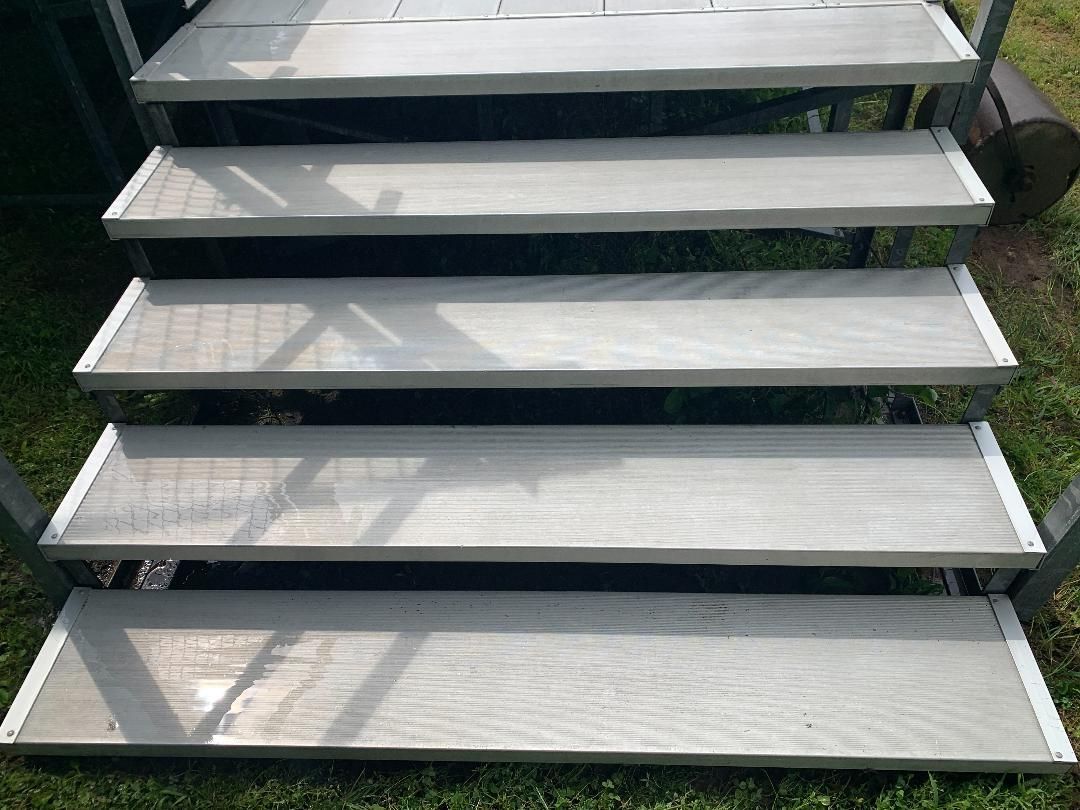 A set of five light gray, wood-grain patterned metal steps leading up, framed by metal side rails outdoors over grass.