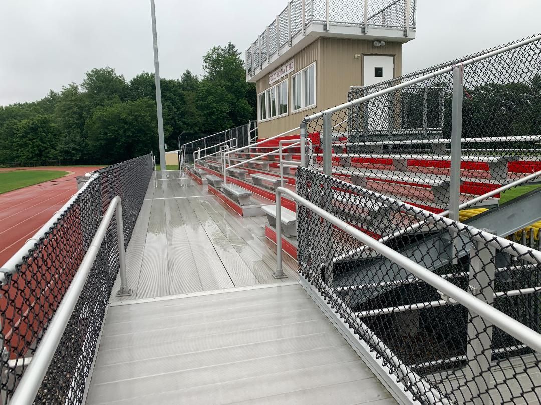 A metal ramp with chain-link railings leads to stadium bleachers next to a running track on a cloudy day.