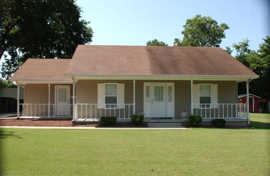 A single-story tan house with a brown roof and a front porch, set against a green lawn and trees.