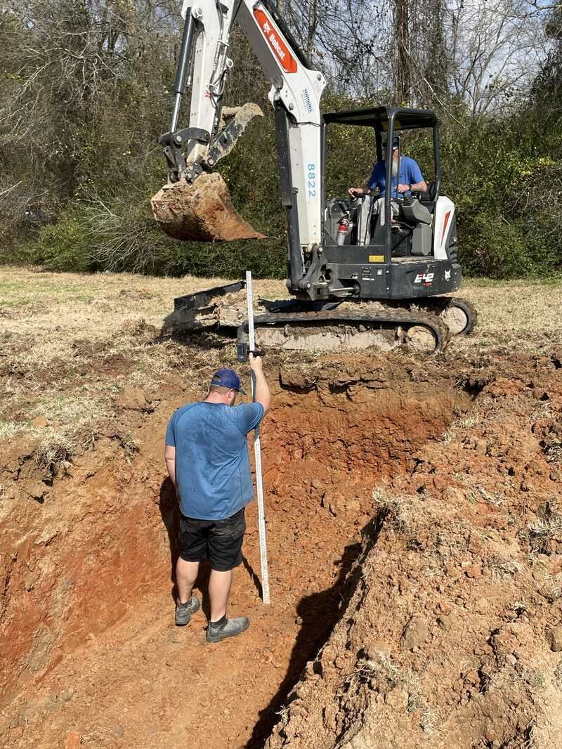 Man using measuring stick as excavator digs trench.