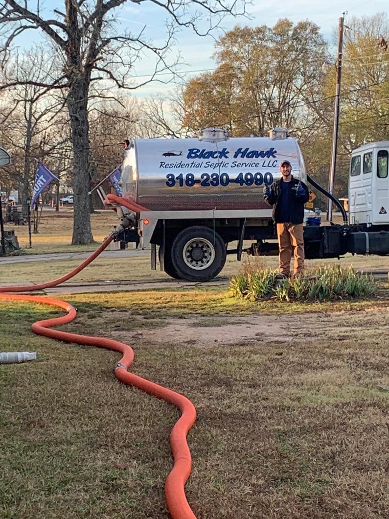 A man stands by a Black Hawk septic service truck with a hose stretched across grass in an outdoor setting.