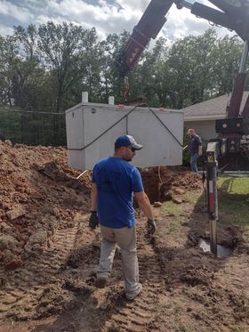 Workers installing a concrete septic tank. Crane lifts tank over an excavated hole in a yard.