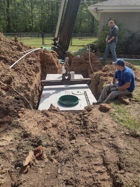 Men installing a septic tank in a trench. A backhoe hoists the tank. Outdoors, daytime.