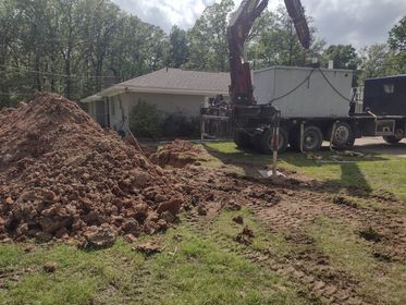 An excavator moving dirt, next to a tank on a truck in front of a house.