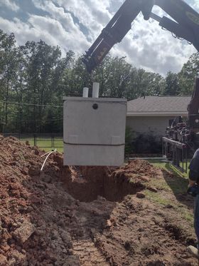 Crane lowering a concrete septic tank into an excavated pit in a yard with trees and a house in the background.
