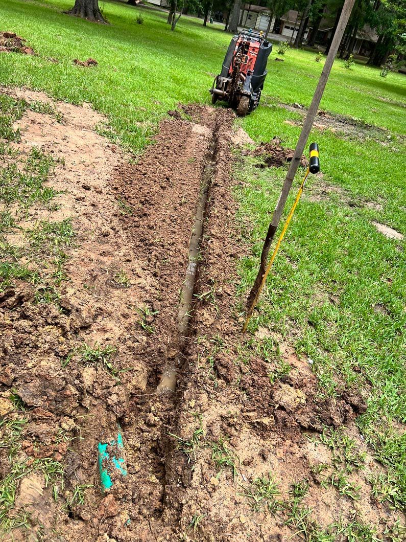 Dug trench in a grassy yard, with a pipe visible. A shovel, lawn machine, and green connectors are also present.