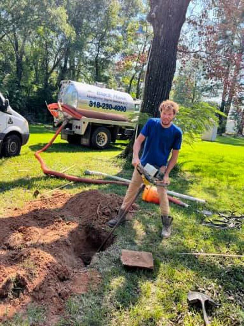Man using a saw near a dug hole, next to a septic truck on a grassy lawn.