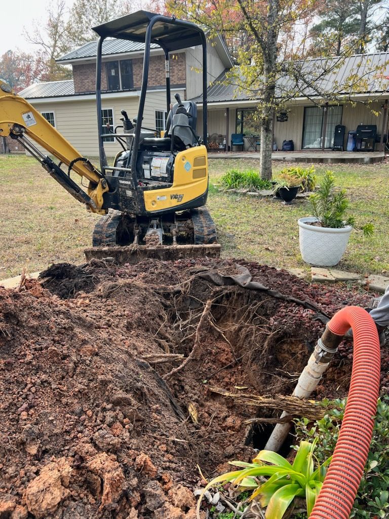 Yellow excavator digging in a yard near a house. Brown dirt piled. Red hose visible.