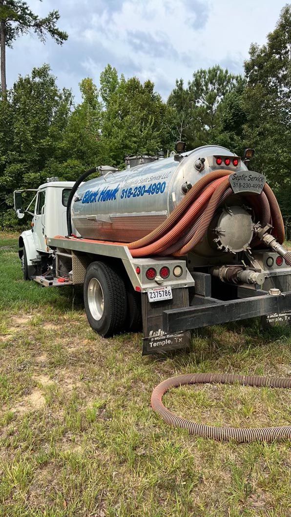 Septic tank service truck parked on grass. Silver tank, orange hose, white cab. Trees in background.