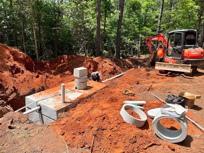 Construction site with a concrete septic tank, backhoe, and red clay soil.