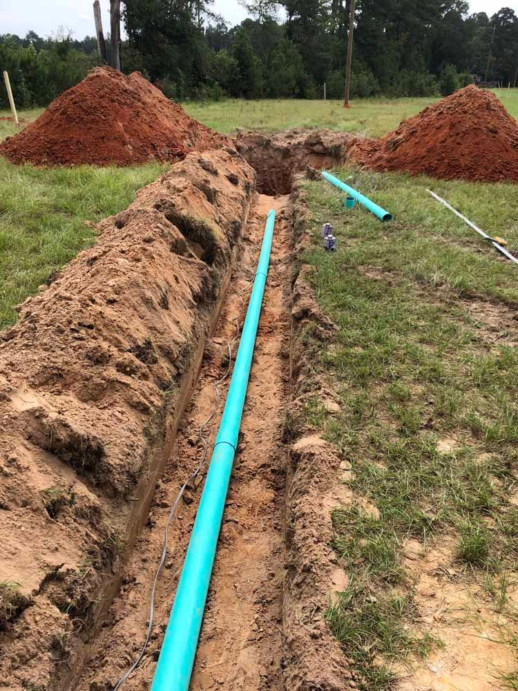 Trench dug in a grassy yard, with blue pipes laid inside; two piles of reddish dirt nearby.