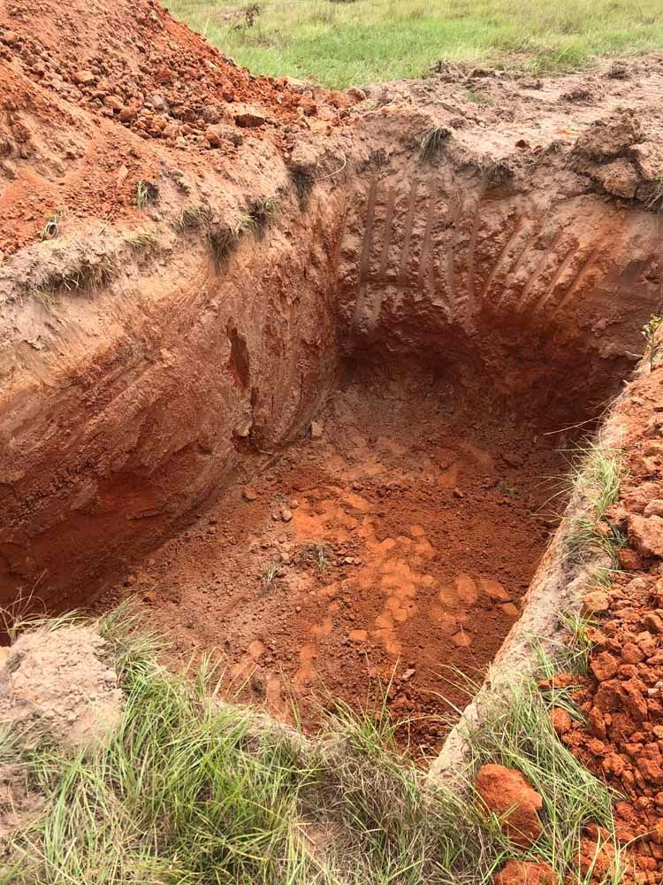 A rectangular hole dug in red soil, with grass along the edge.