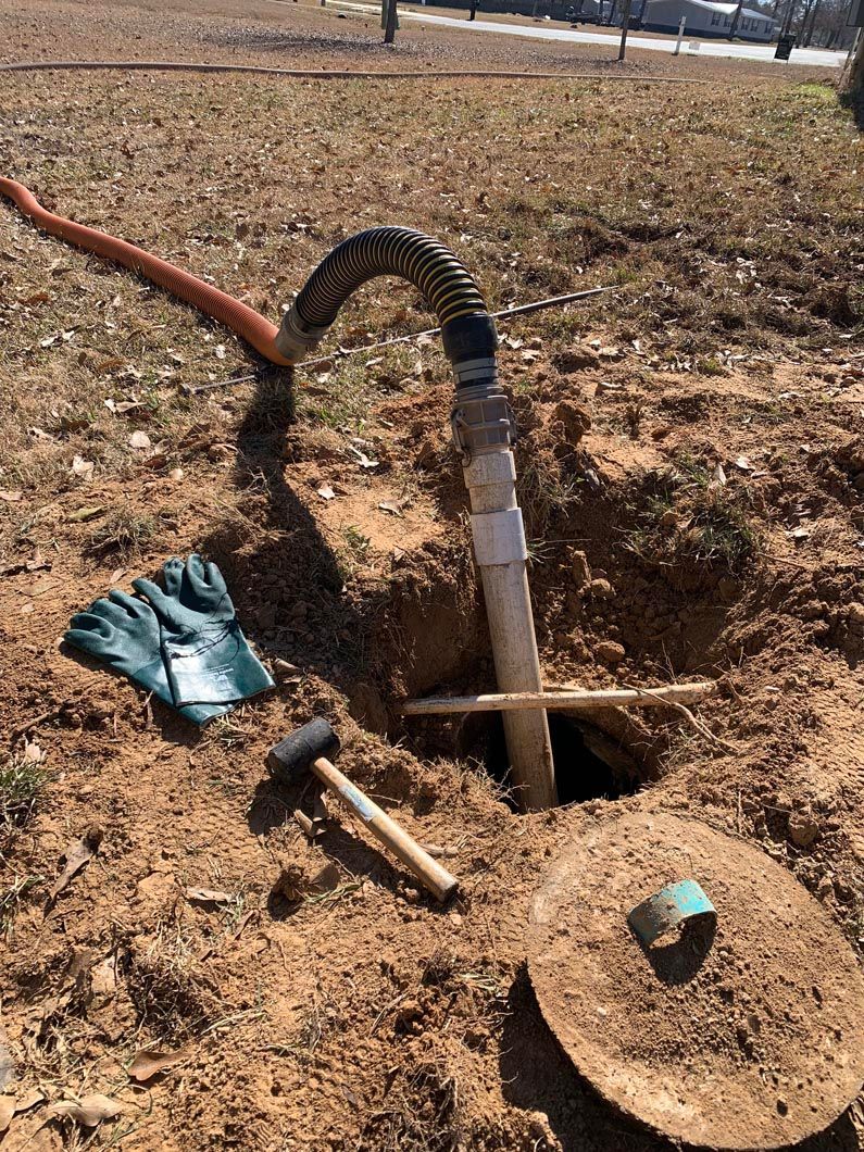 Hole in the ground with a pipe and hose. Tools and a lid sit nearby, implying septic tank maintenance.