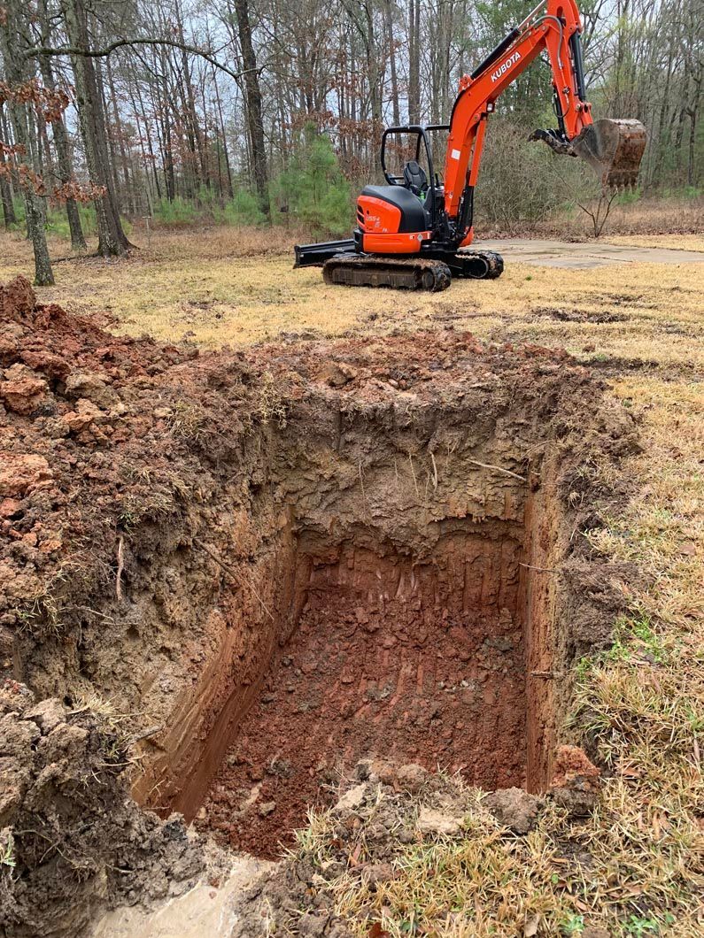 Mini excavator digging a square hole in a field; orange machine, dirt, trees in background.