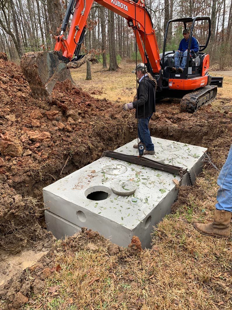 Workers installing a concrete septic tank using an excavator in an outdoor setting.