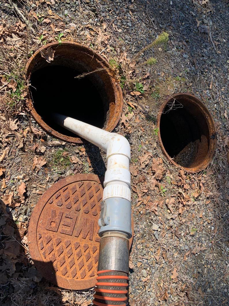 A septic tank being pumped. A white hose extends into an open tank, lid open nearby, set outdoors.