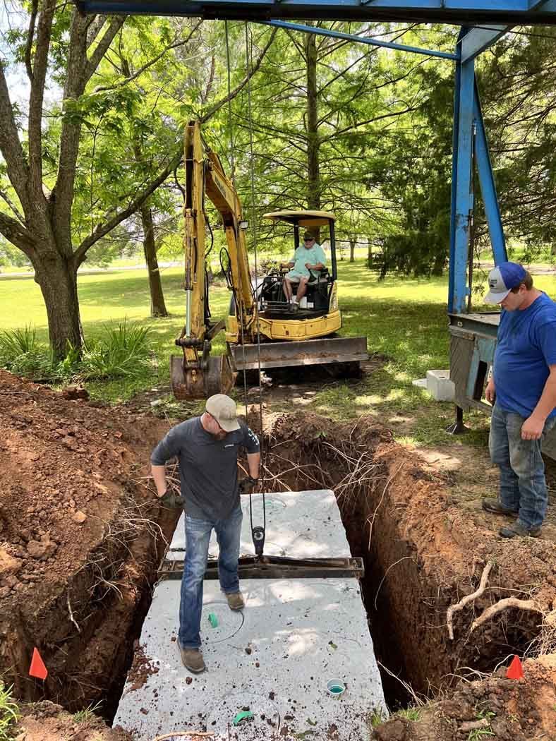 Men installing a septic tank with an excavator in a grassy yard, under a blue frame.