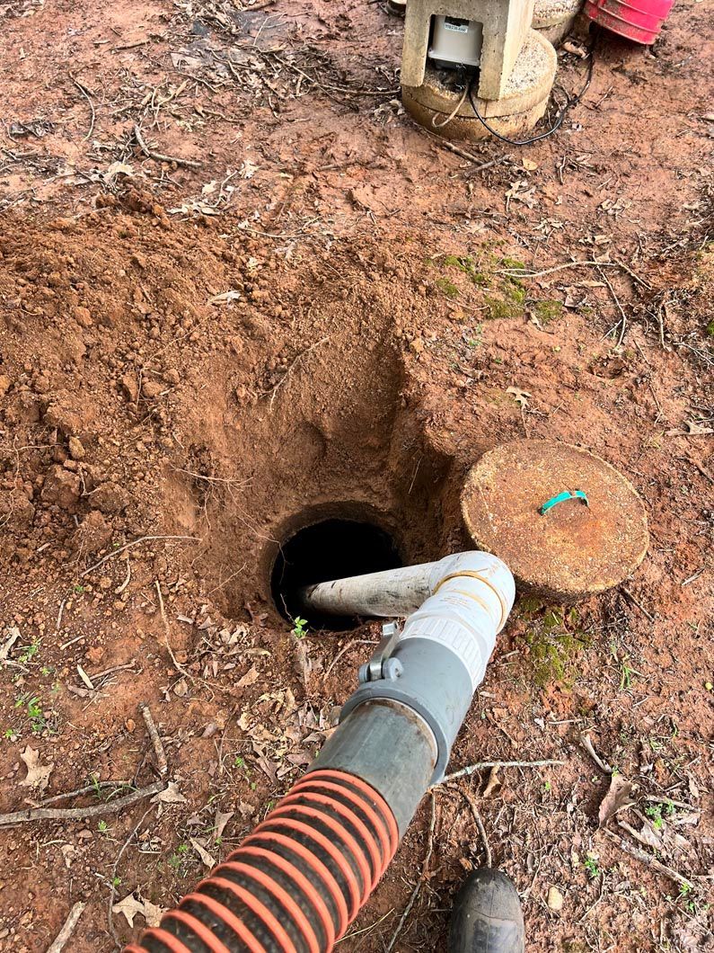 Hole dug in dirt with open septic tank, hose inserted. Orange and gray hose with round cover.