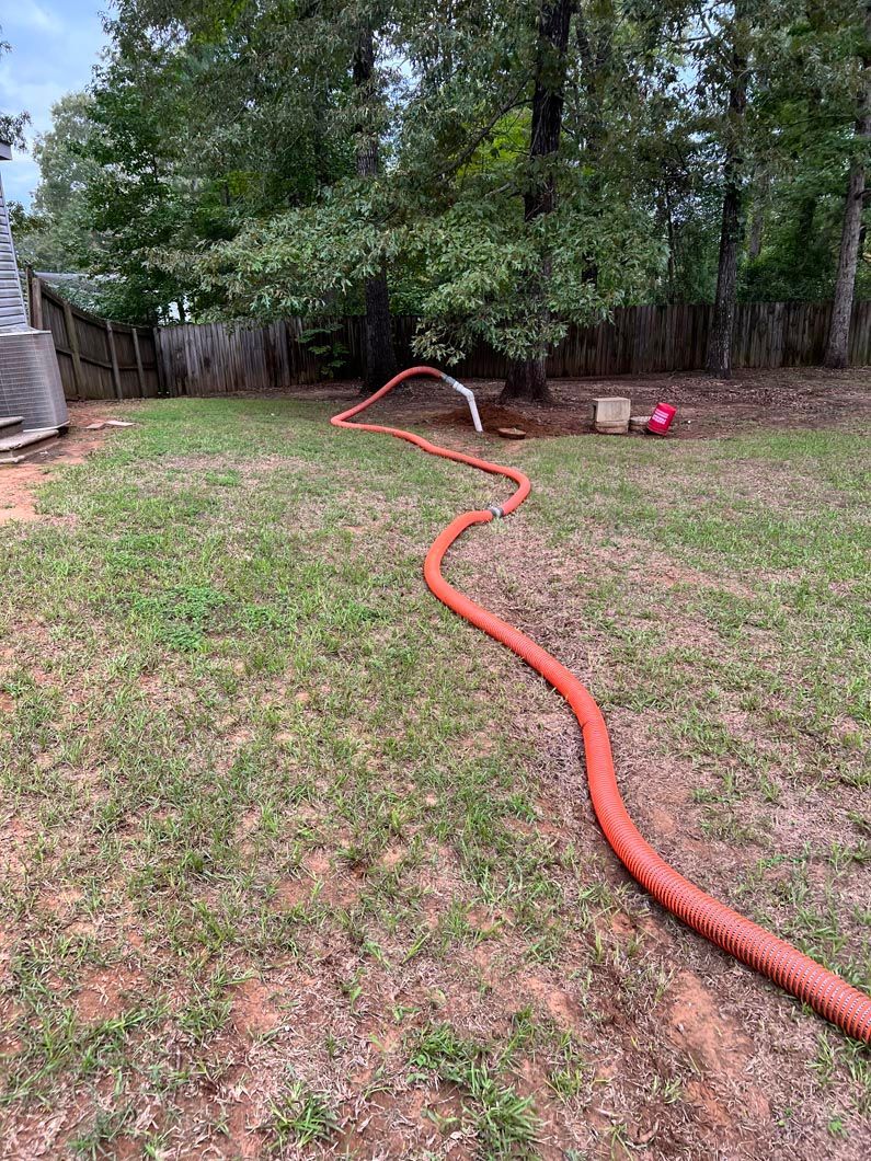 Orange hose spraying water in a grassy backyard with trees and a wooden fence.