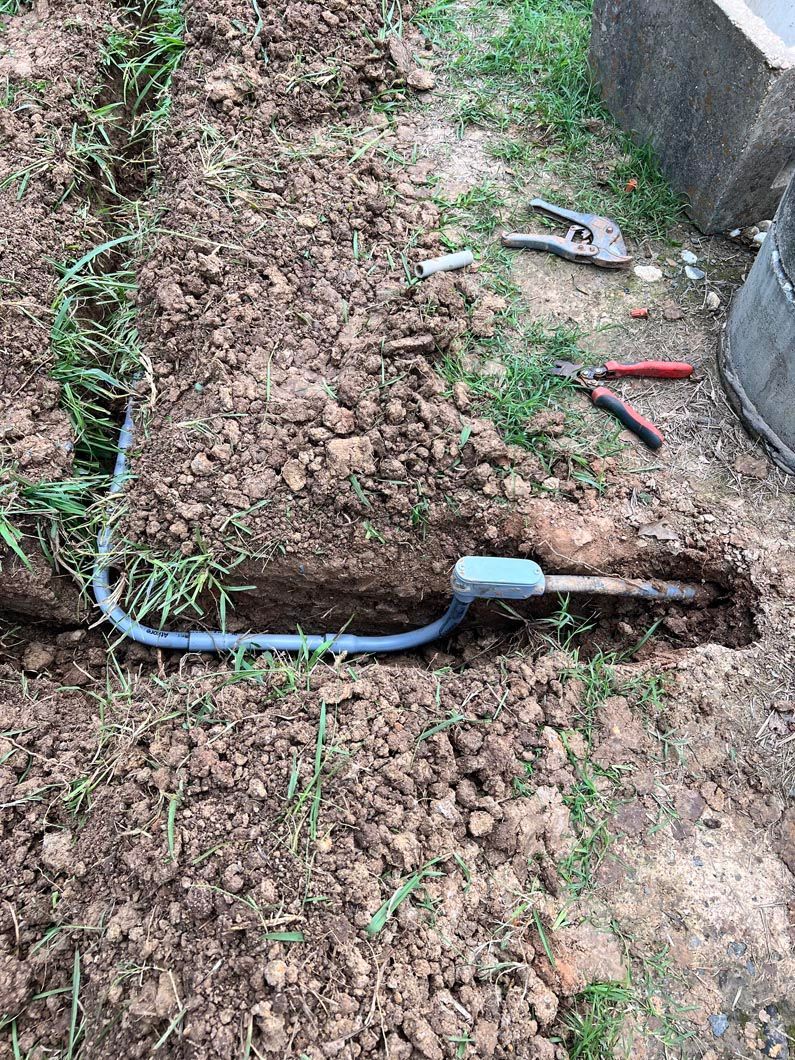 Trench with electrical conduit and junction box. Tools on ground. Outdoor setting, brown soil, green grass.